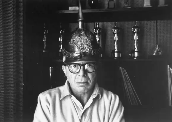 Director  Billy  Wilder  in  his  Hollywood  office  wearing  German  helmet  w.  Oscars  on  shelf